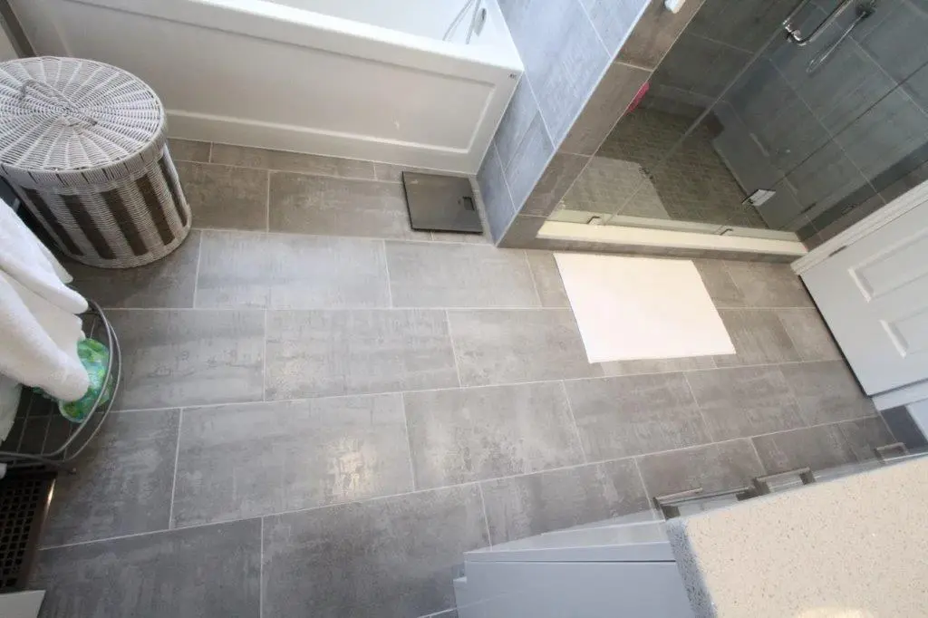 A modern bathroom with gray tiled floors and shower walls, featuring a glass shower enclosure, a white bath mat, a wicker laundry hamper, and a stack of white towels.