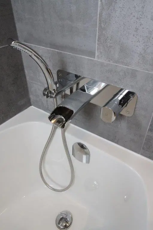 A modern chrome bathroom faucet with a handheld shower attachment above a white bathtub, set against a gray tiled wall.
