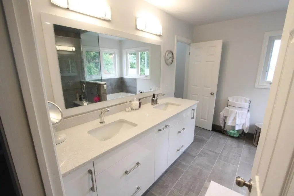 A modern bathroom with a dual-sink white vanity under a large mirror, featuring overhead lighting. The room has gray tiled flooring, a shower with a glass door, and windows allowing natural light. There is a clock on the wall and a towel rack next to the door.