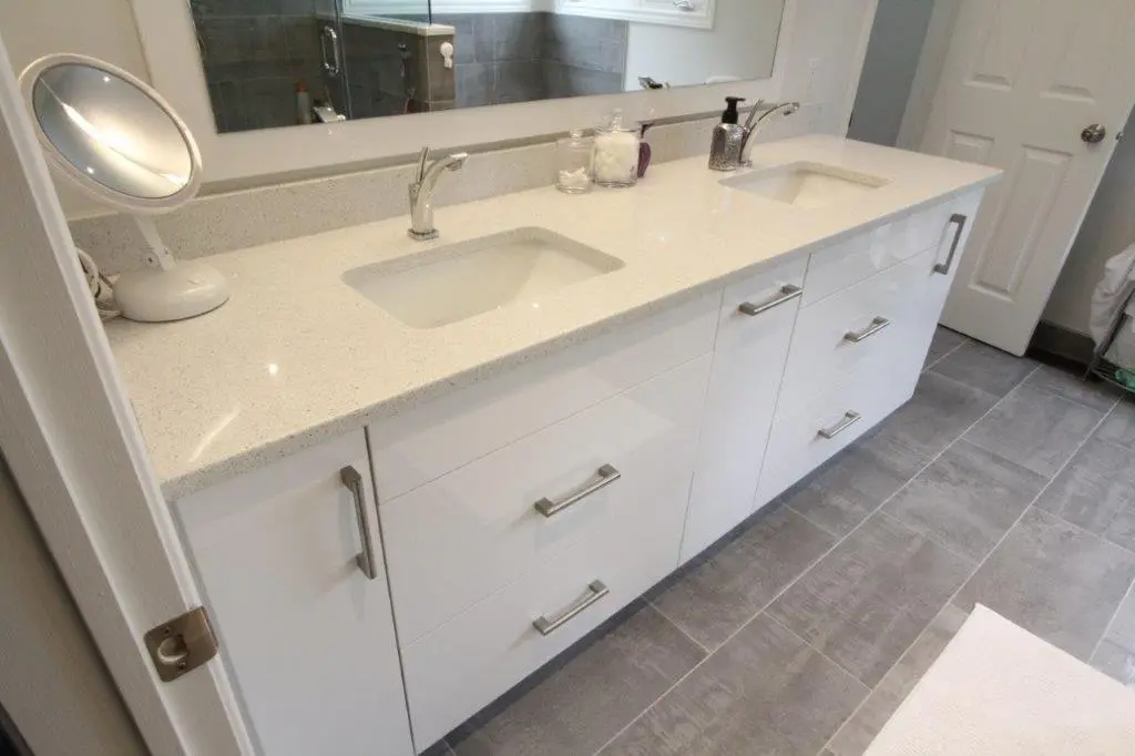 A bathroom vanity with a light-colored countertop featuring two built-in sinks, faucets, and a makeup mirror. The cabinet below has white drawers with metallic handles, and there's a soap dispenser and jar on the countertop. The floor is covered with gray tiles.