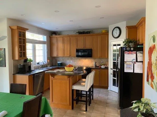 A before renovation kitchen with wooden cabinets, a kitchen island with two stools, a large window, and a black refrigerator. The counters host a coffee maker and fruit bowl, and there's a clock on the wall next to a door with glass panels. A dining table with a green tablecloth is partially visible.