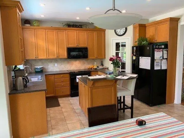 A before renovation kitchen with wooden cabinets, black appliances including a stove and refrigerator, a center island with chairs, and a table with a striped tablecloth in the foreground.