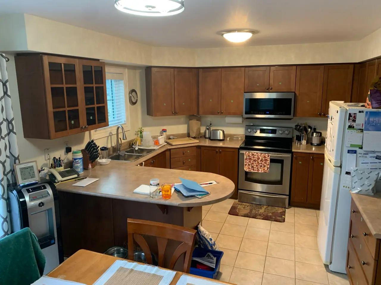 A before renovation kitchen with wooden cabinets, a stainless steel stove, a microwave, and a double sink. The counter has various items, including paper towels, cleaning supplies, and kitchen utensils. There is a refrigerator on the right side, and a dining table with a chair in the foreground.