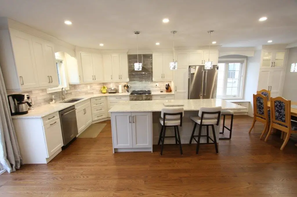 A modern kitchen with white cabinets, a large quartz island with three chairs, stainless steel appliances, and a dining area with wooden chairs and a table.