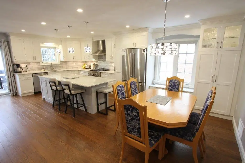 A modern kitchen with white cabinets, an island with bar stools, and a wooden dining table with upholstered chairs placed on a hardwood floor.