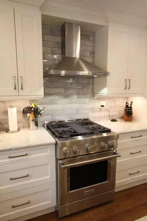 A modern kitchen with white cabinets features a stainless steel gas range and hood, with a gray subway tile backsplash and a quartz countertop. A knife block, utensil holder, and paper towel stand are on the counter.