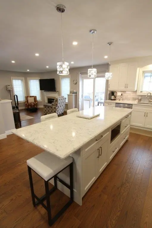 A modern kitchen with a large quartz island featuring bar stools, surrounded by hardwood floors, white cabinetry, and hanging pendant lights. The space opens into a cozy living area with a fireplace and a wall-mounted TV.