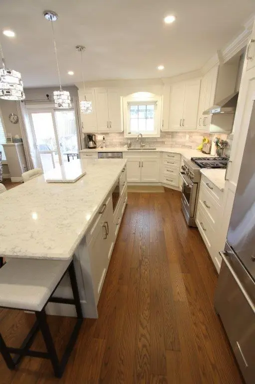 A modern kitchen with white cabinetry, quartz countertops, and stainless steel appliances. The room features a central island with seating, hardwood floors, and pendant lighting.