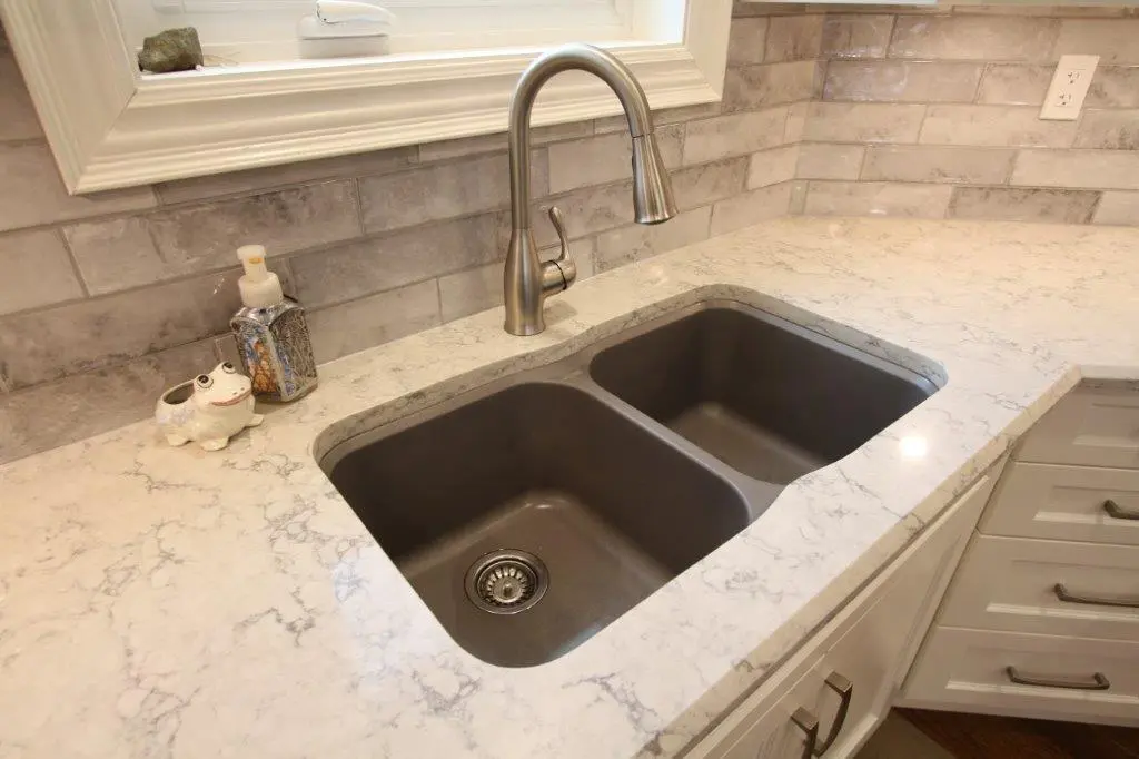 A kitchen sink with a dual-basin setup and a stainless steel faucet, set in a white quartz countertop. The backsplash is made of light gray tiles. There is a soap dispenser and a ceramic frog decoration on the counter.