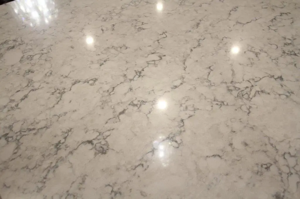Close-up of a polished quartz countertop with a light gray and white veined pattern, reflecting light from overhead fixtures.