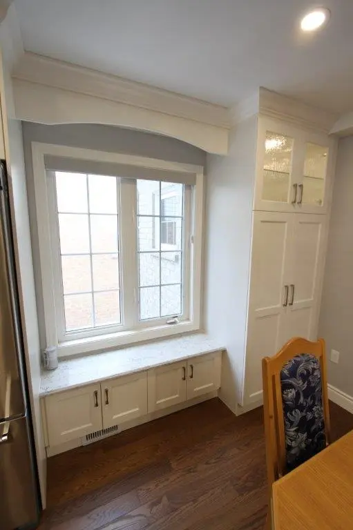 A kitchen corner with a large window, white cabinetry with glass-paneled doors, a small seating area with a cushion below the window, and a wooden chair with a patterned cushion next to a wooden table on a hardwood floor.