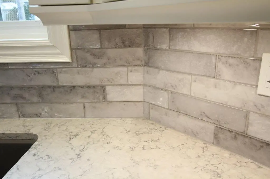 A quartz kitchen countertop with a marble-like surface and a gray subway tile backsplash, featuring a window and a portion of a white cabinet above.
