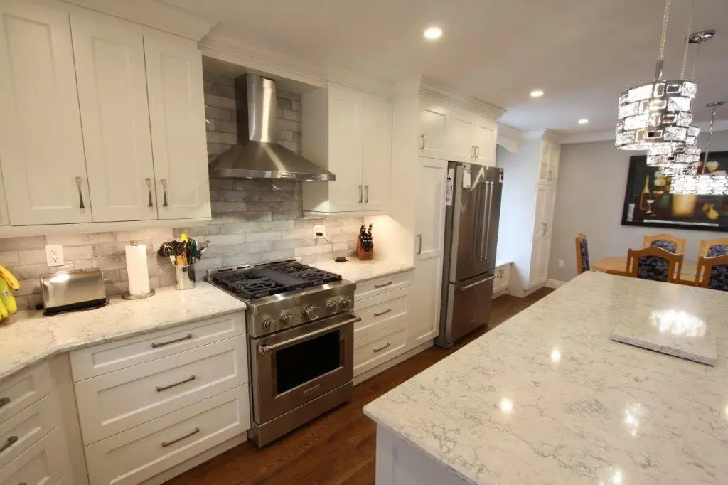 Modern kitchen with white cabinets, stainless steel appliances, and quartz countertops, featuring a central island with decorative pendant lighting and a dining area in the background.