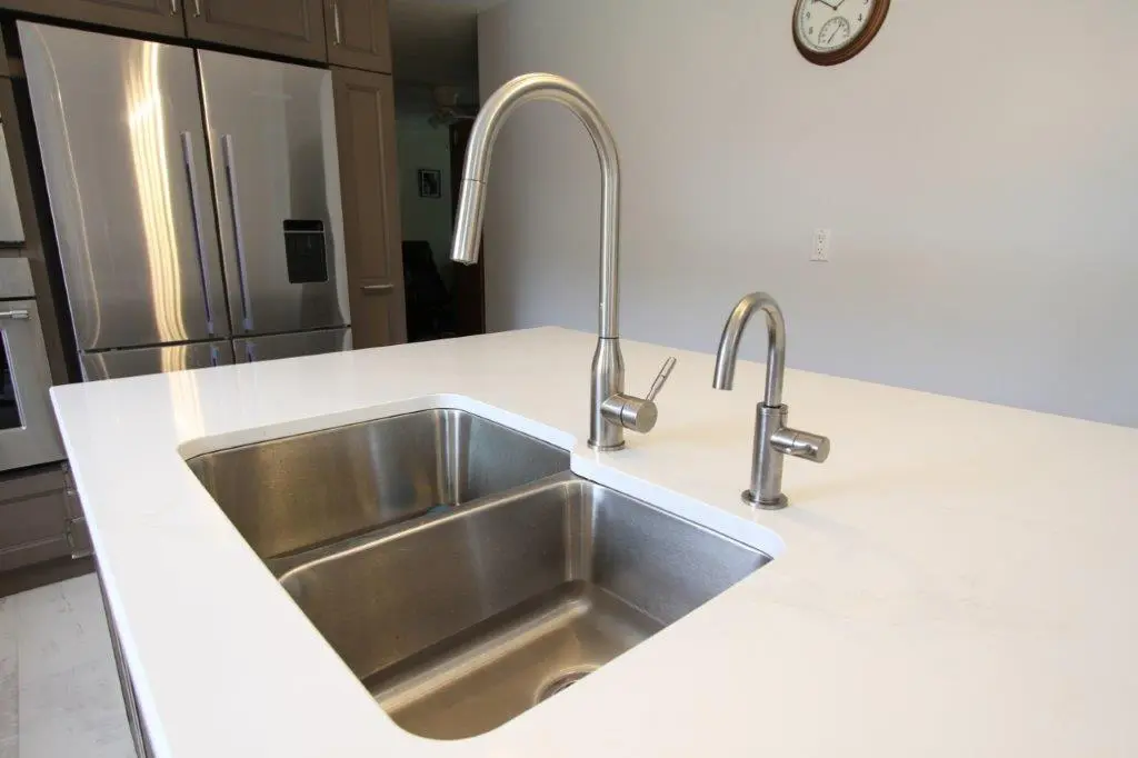 Modern kitchen with a stainless steel sink and faucet set into a white quartz countertop, with stainless steel appliances in the background.