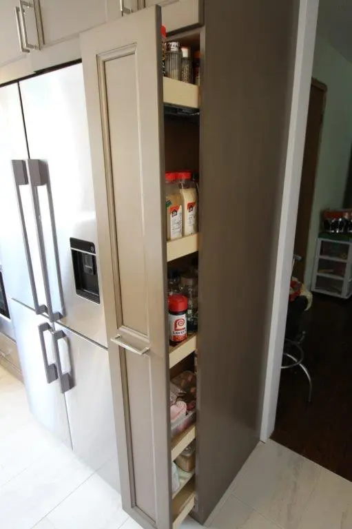 A narrow pull-out pantry next to a stainless steel refrigerator, containing shelves filled with various spice jars and bottles.