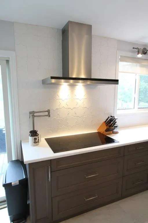 Modern kitchen with a sleek black induction cooktop, stainless steel range hood, and patterned white tile backsplash. There is a wooden knife block and a jar beside the stove, and cabinets in a dark wood finish below.