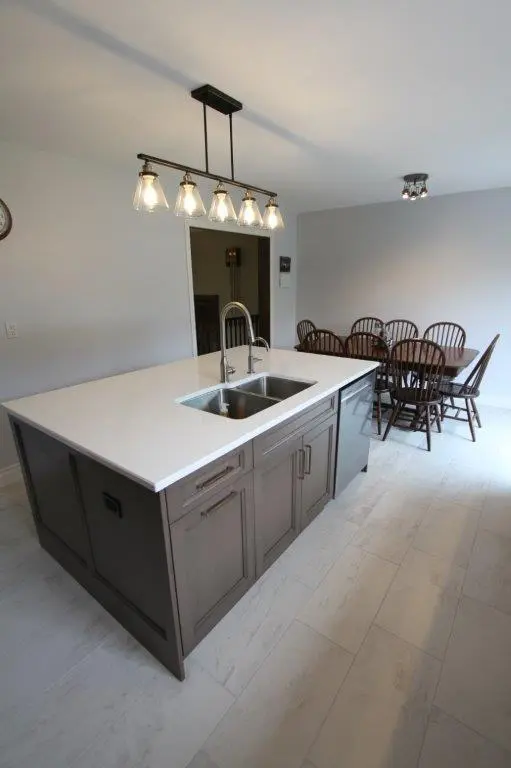 Modern kitchen with a large island featuring a white quartz countertop and stainless steel sink, illuminated by a stylish hanging light fixture, with a dining area in the background including a wooden table and chairs.