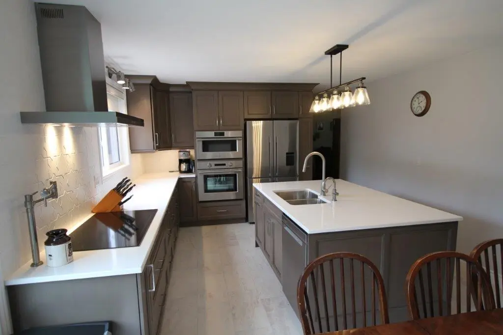 A modern kitchen with brown cabinetry, stainless steel double ovens, a large refrigerator, and an island with a sink. It features a cooktop on the left countertop and overhead pendant lights above the island.