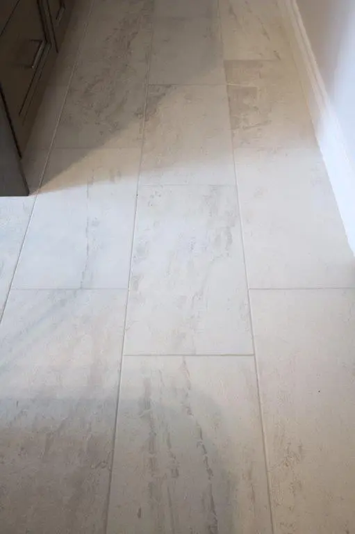 A floor with large, rectangular beige tiles, showing subtle variations in color and texture, adjacent to dark cabinetry.