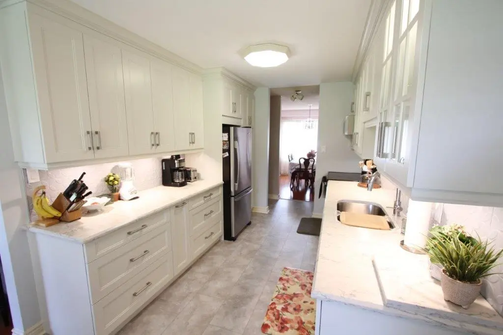 A modern kitchen with white cabinetry, quartz countertops, and stainless steel appliances. There are decorative plants, a knife block, and small kitchen appliances on the counters. A dining area is visible through a doorway.