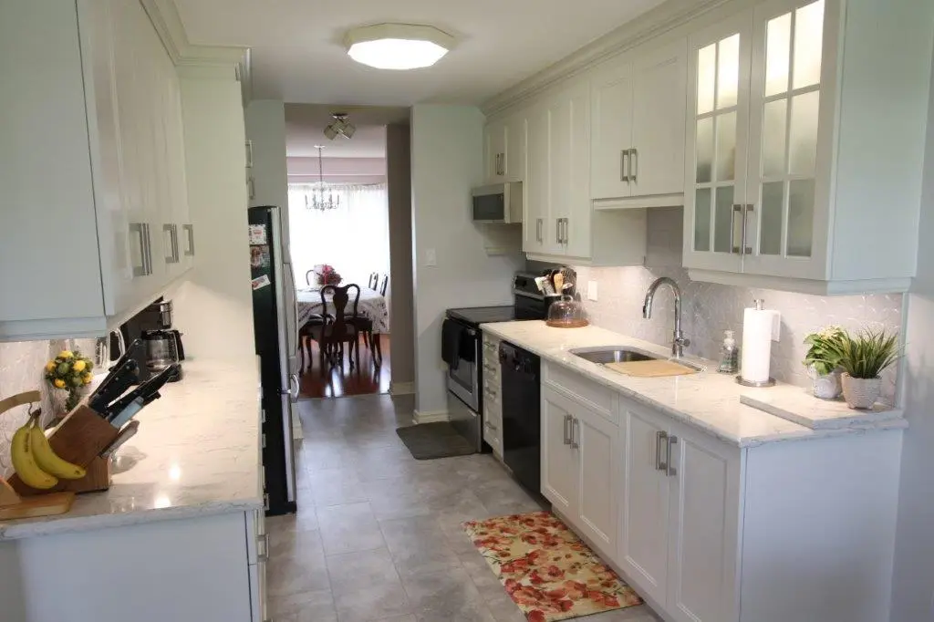 Bright, narrow kitchen with white cabinets, quartz countertops, and stainless steel appliances, leading into a dining area.