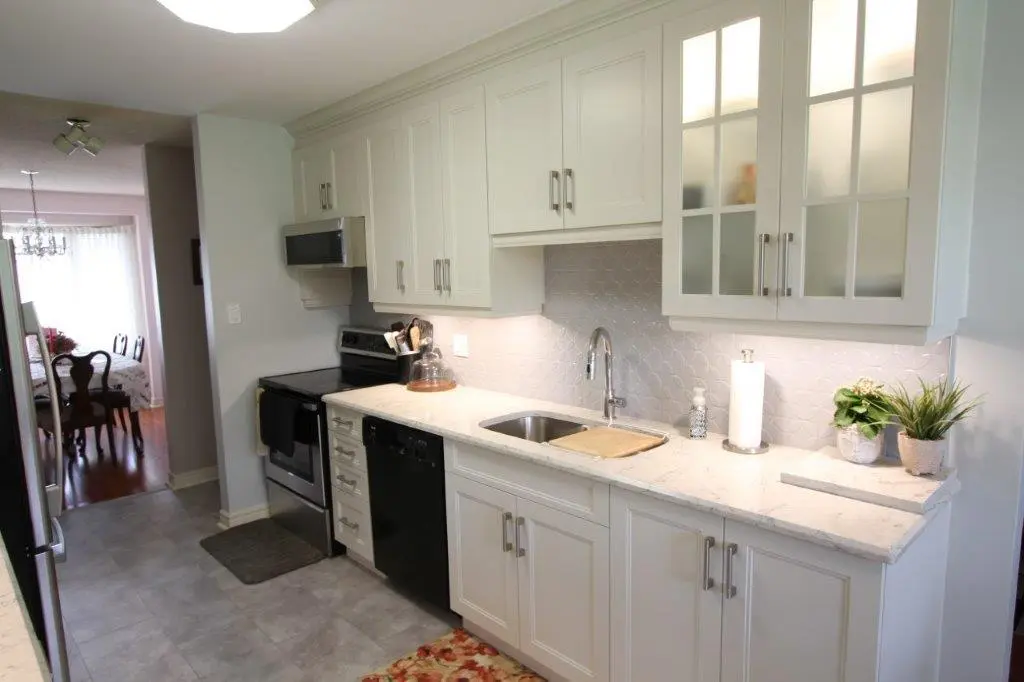A modern kitchen with white cabinets, a stainless steel stove, and a sink with a metallic faucet. The backsplash features a light grey pattern, and the countertops are white with decorative plants. A dining area is visible through a doorway.