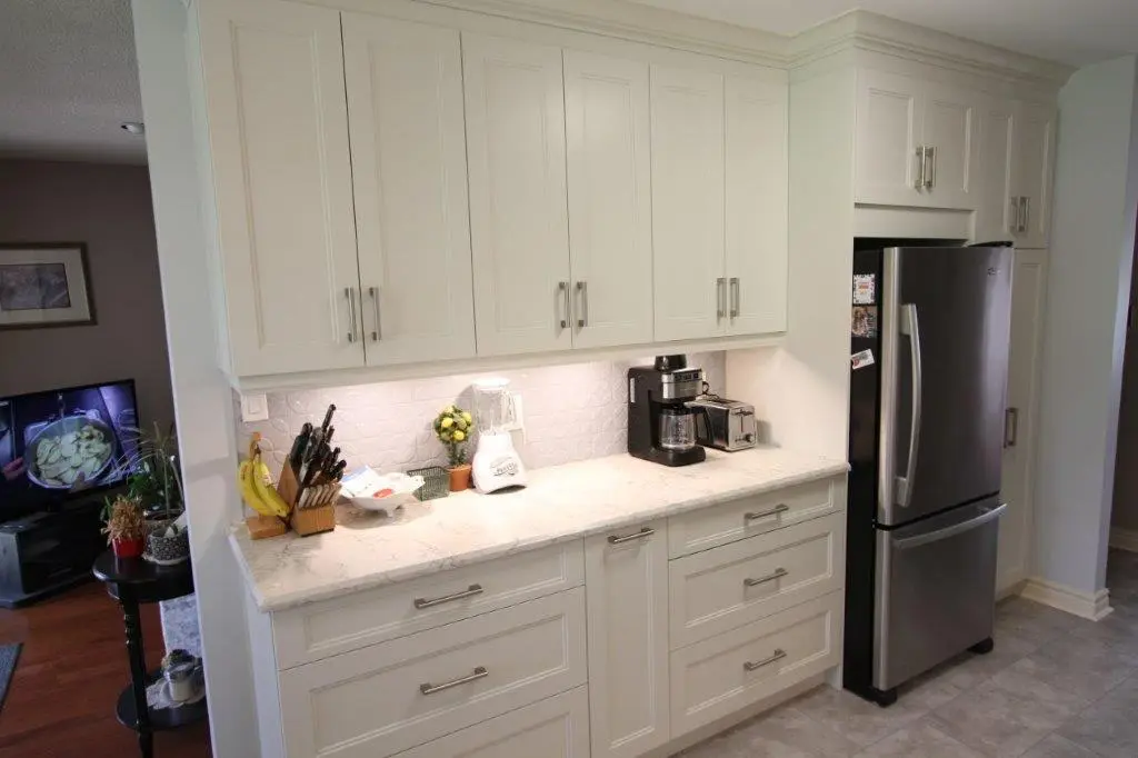 A modern kitchen with white cabinets and drawers, featuring a quartz countertop. On the counter, there is a knife block, a bowl of bananas, a blender, and a coffee maker. A stainless steel refrigerator is installed. The backsplash is made of textured white tiles, and some decorative objects are placed on the counter.