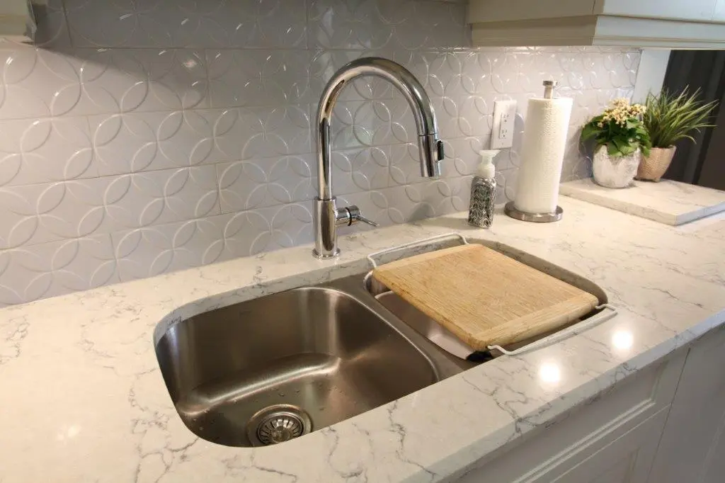 A modern kitchen sink with a shiny silver faucet mounted on a marble countertop. A wooden cutting board rests over one section of the sink. The backsplash features a geometric circular pattern, and there is a soap dispenser, a roll of paper towels, and a small potted plant nearby.