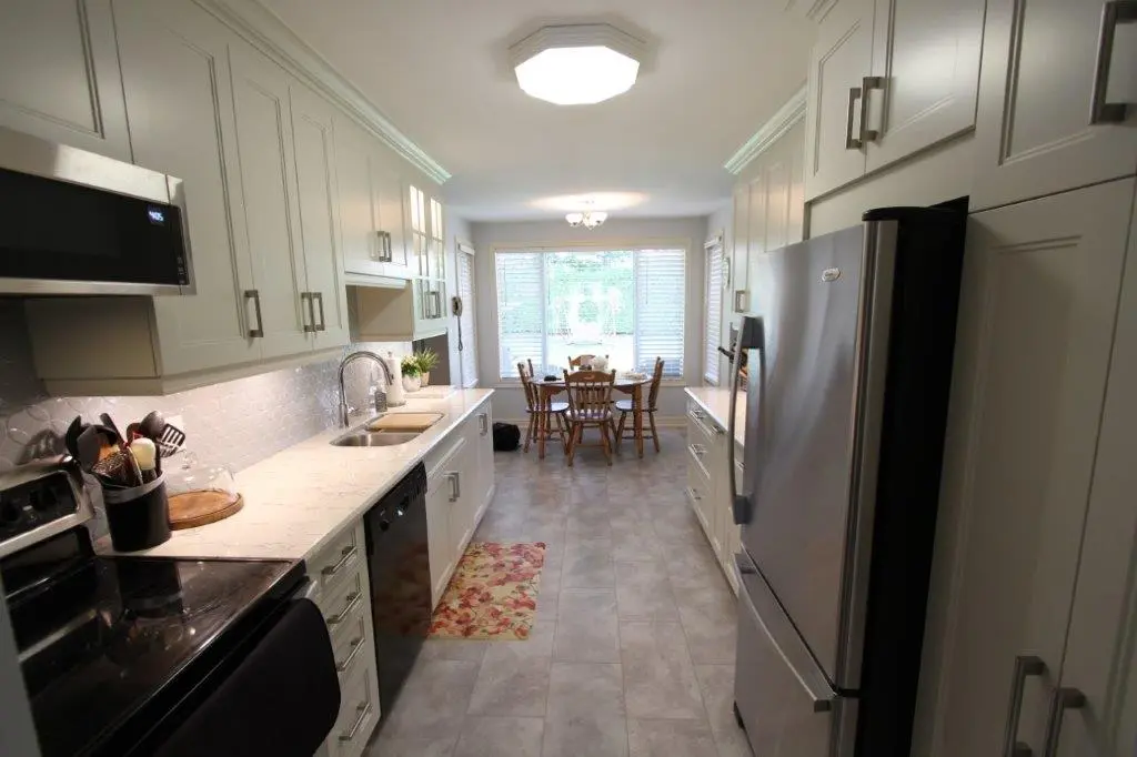A modern galley kitchen with white cabinets, stainless steel appliances, and a quartz countertop featuring a sink. The kitchen leads to a dining area with a wooden table and chairs near a large window with blinds. The floor is light grey tile, and a floral rug is placed in front of the sink.