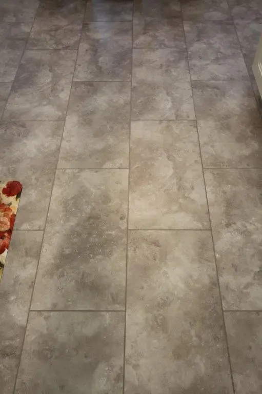 A floor with large rectangular gray tiles featuring a subtle marbled pattern, with a partial view of a floral patterned rug on the left edge.