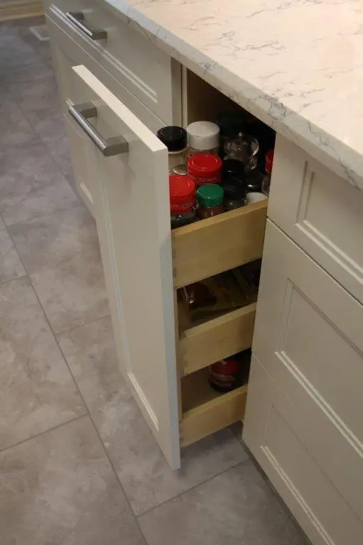 A kitchen cabinet with a pull-out spice rack containing various spice jars, positioned beneath a quartz countertop.