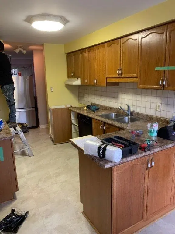 A "before" kitchen under renovation with wooden cabinets, a double sink on the countertop. The walls are painted yellow, and the floor is tiled.