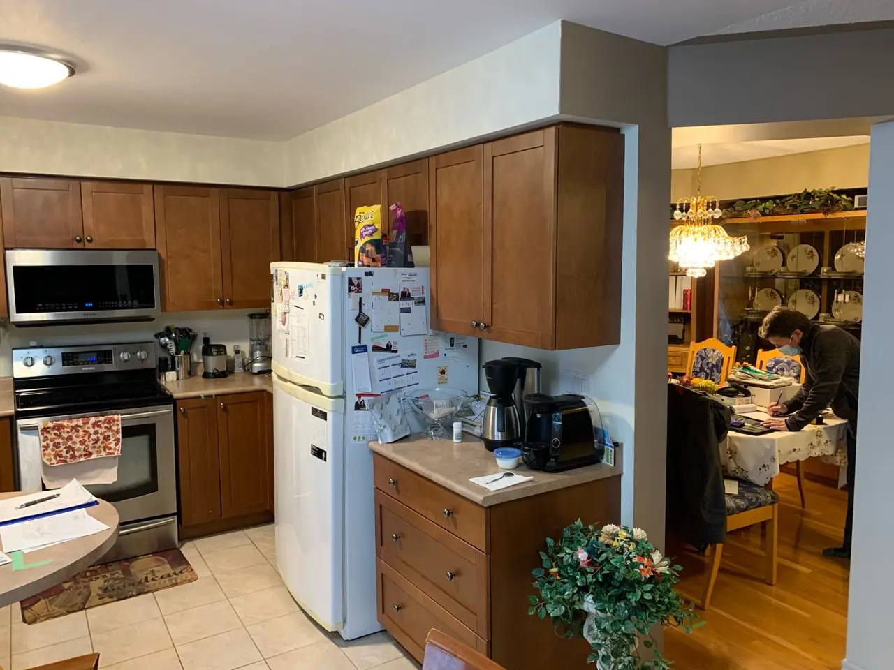 A before renovation kitchen with wooden cabinets, a white refrigerator covered with magnets and papers, and stainless steel appliances.