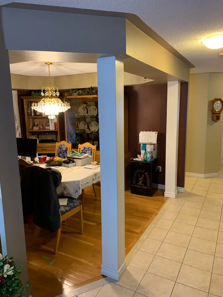 A before renovation dining area with wooden flooring, a rectangular table covered with a tablecloth, and chairs with blue patterned cushions. A chandelier hangs above the table. A hutch displaying plates and a clock on the wall are visible. The dining space is adjacent to a tiled hallway.
