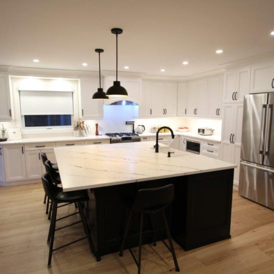 A modern kitchen with white cabinets, a large central island featuring a quartz countertop, black stools, and a black faucet. Stainless steel appliances include a refrigerator and stove, with overhead pendant lighting and wooden flooring.