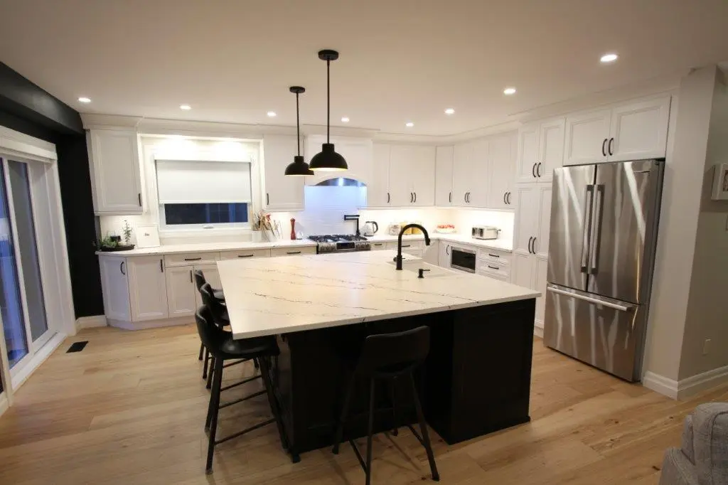 A modern kitchen with white cabinets, a large central island featuring a quartz countertop, black stools, and a black faucet. Stainless steel appliances include a refrigerator and stove, with overhead pendant lighting and wooden flooring.