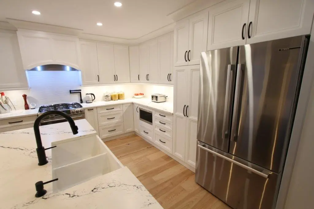A modern kitchen with white cabinetry, a large stainless steel refrigerator, farmhouse sink, and light wood flooring. The countertops are white with black veining, and there are various small appliances and utensils on the counters.