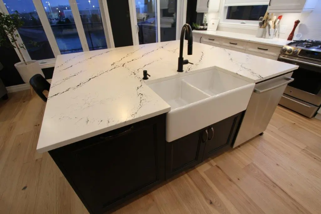 A modern kitchen island featuring a white quartz countertop with black veining, a white farmhouse sink, a black faucet, and dark cabinetry. The floor is light wood, and large windows in the background provide natural lighting.