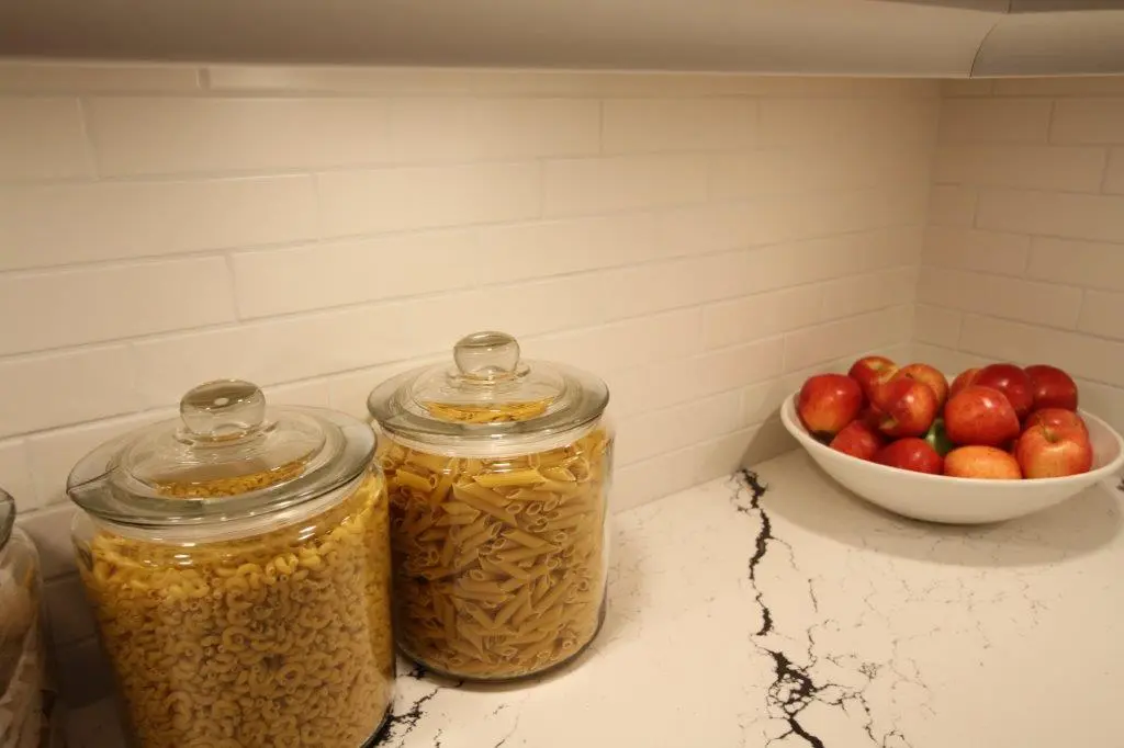 A quartz kitchen counter with two glass jars filled with dry pasta, and a white bowl holding several red apples, set against a white tiled backsplash.