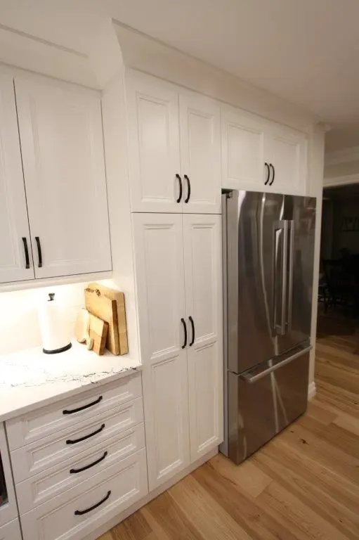 Modern kitchen with white cabinets and a stainless steel refrigerator, featuring a wooden floor, black handles, and a countertop with cutting boards and a paper towel holder.