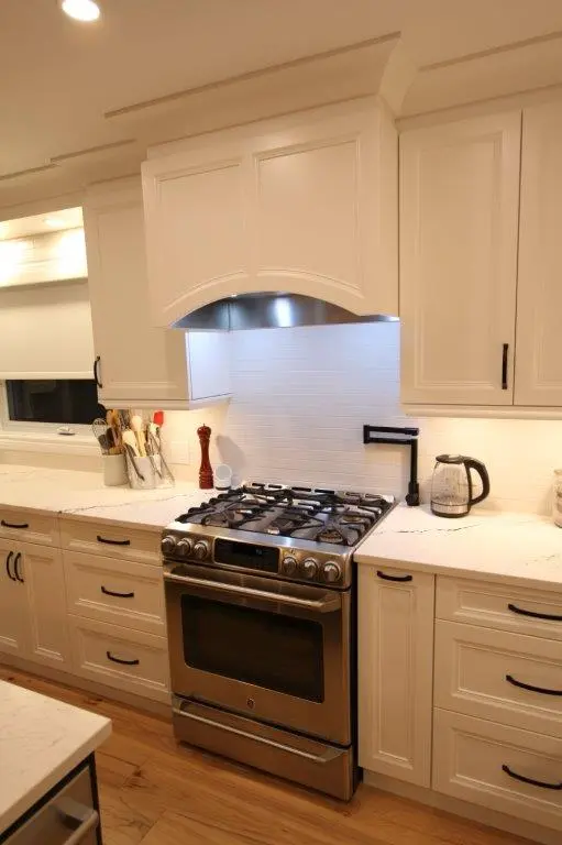 Modern kitchen with a stainless steel oven and gas stove, surrounded by white cabinets and a quartz countertop, featuring cooking utensils, a pepper grinder, and an electric kettle on the counter.
