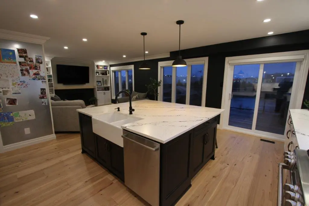 Spacious modern kitchen with a large quartz-topped island featuring a farmhouse sink and pendant lights above, adjacent to a living area with a flat-screen TV and sliding glass doors leading to an outside deck.