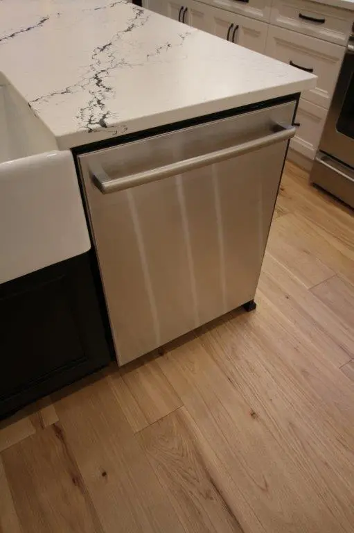 A modern kitchen with a stainless steel dishwasher integrated into a wooden cabinet, featuring a light wood floor and a white quartz countertop with dark veining.