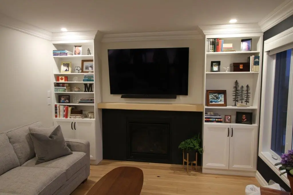 A living room with a large TV mounted above a fireplace, flanked by white built-in shelves filled with books, photos, and decorative items. A gray sofa is positioned in front of a wooden coffee table, and a window is visible on the right side of the image.