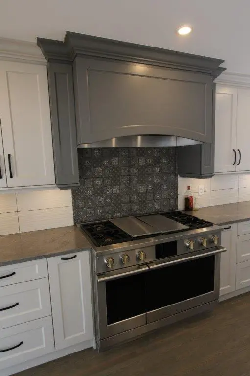 A modern kitchen stove with a stainless steel finish, flanked by white cabinetry and featuring a decorative tiled backsplash under a large, gray range hood.