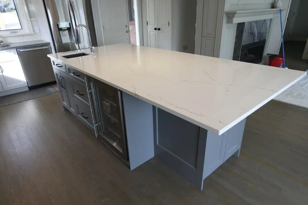 A modern kitchen island with a smooth, white countertop and gray cabinetry, equipped with a sink, integrated appliances, and a wine cooler, set on a wooden floor with a stainless steel refrigerator in the background.