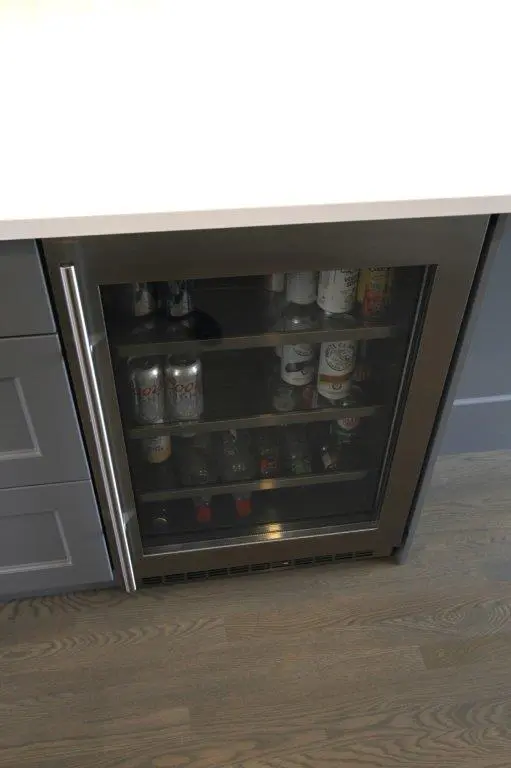 A beverage refrigerator with a glass door, containing various cans and bottles, integrated into a kitchen cabinet.