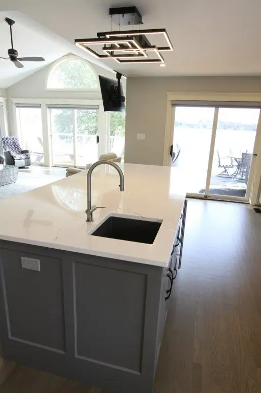 Modern kitchen interior featuring a large white quartz island with a sink, contemporary ceiling light fixture, and a view of a living area with large windows and sliding glass doors leading to an outdoor patio with a scenic lake view.