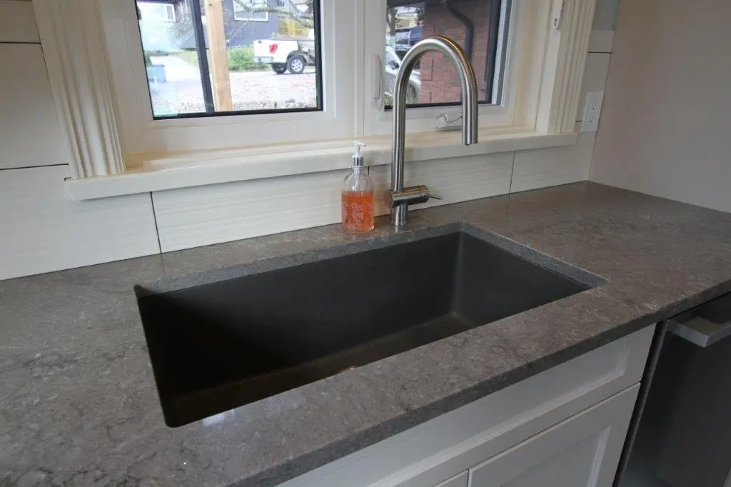A modern kitchen sink with a sleek, dark basin and a stainless steel faucet, set in a gray quartz countertop, with a window and a bottle of liquid soap nearby.