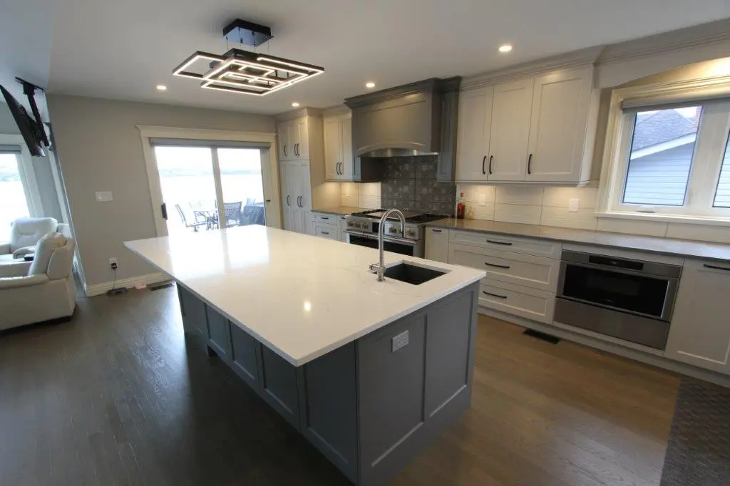 Modern kitchen with a large white island countertop, pendant lighting, grey cabinets, stainless steel appliances, and sliding glass doors leading to an outdoor patio.